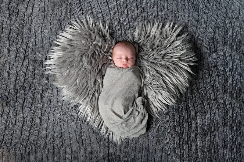 Color photo of a newborn baby boy in a grey wrap on a grey heart rug on a grey backdrop. The photo is by Laura Pea Photography, a Long Island newborn and pet photographer. 