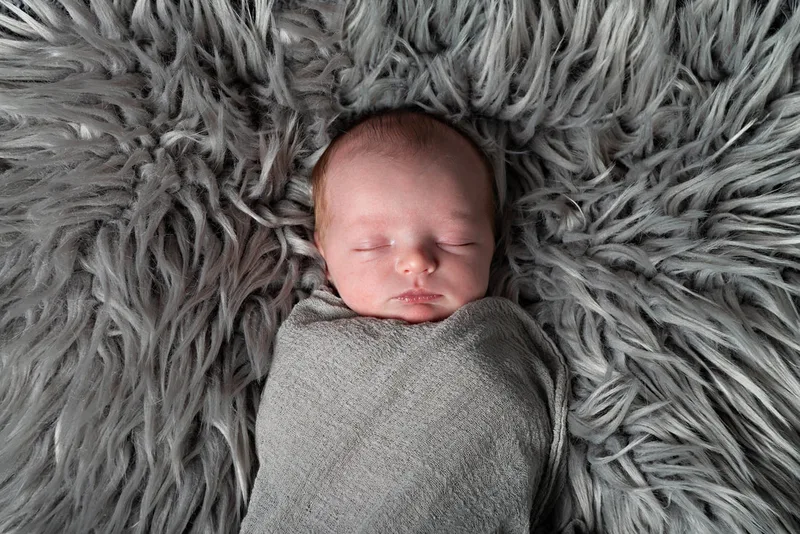 Color photo of a newborn baby boy in a grey wrap on a grey rug. The photo is by Laura Pea Photography, a Long Island newborn and pet photographer. 