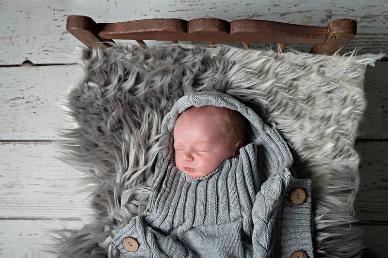 Color photo of a newborn baby boy in a grey knit sack on a vintage doll bed. The photo is by Laura Pea Photography, a Long Island newborn and pet photographer. 