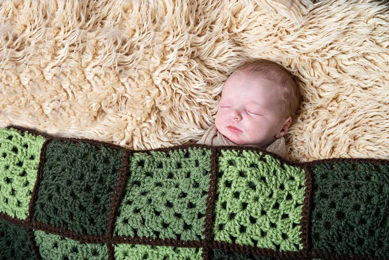 Color photo of a newborn baby boy under a handmade green quilt on a beige rug. The photo is by Laura Pea Photography, a Long Island newborn and pet photographer. 
