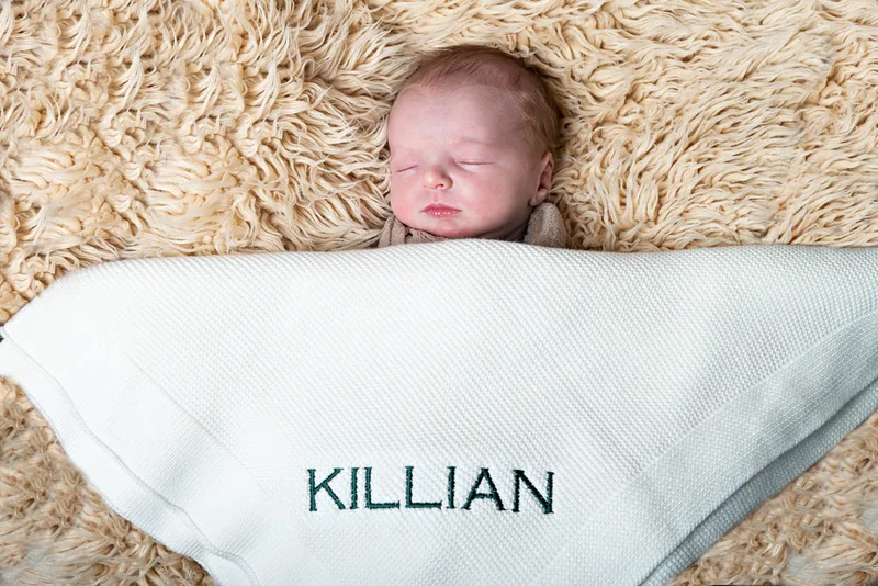Color photo of a newborn baby boy under a blanket that reads "Killian", his name. The photo is by Laura Pea Photography, a Long Island newborn and pet photographer. 