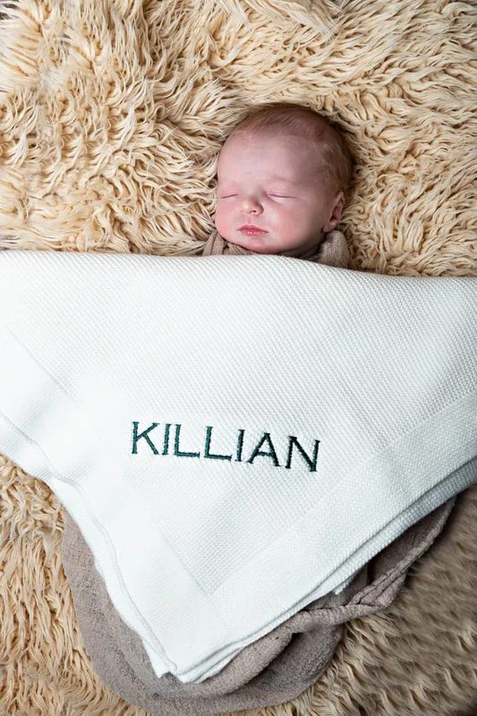 Color photo of a newborn baby boy under a blanket that reads "Killian", his name. The photo is by Laura Pea Photography, a Long Island newborn and pet photographer. 