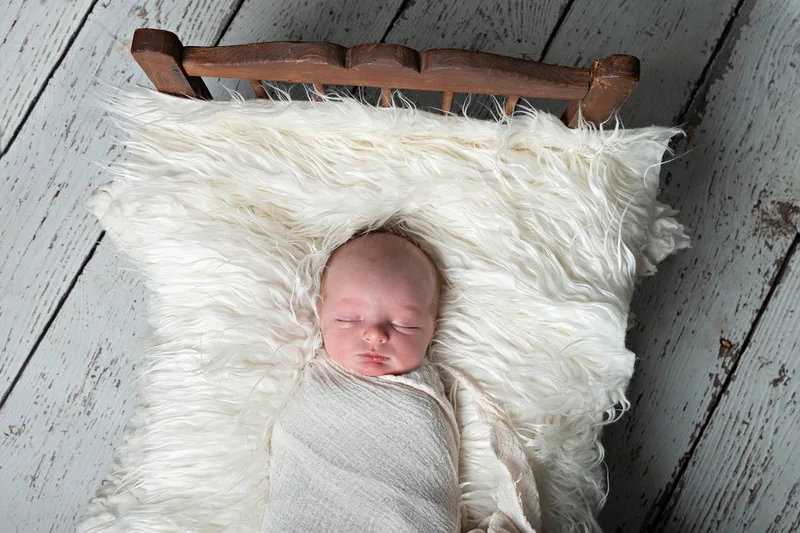 Color photo of a newborn baby boy in a white wrap on a vintage doll bed. The photo is by Laura Pea Photography, a Long Island newborn and pet photographer. 