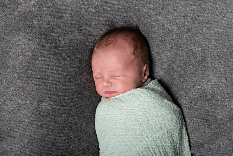 Color photo of a newborn baby boy in a green wrap against a grey fabric backdrop. The photo is by Laura Pea Photography, a Long Island newborn and pet photographer. 