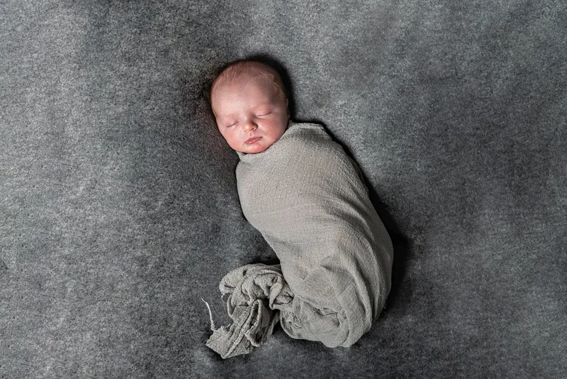 Color photo of a newborn baby boy in a beige wrap on a grey backdrop. The photo is by Laura Pea Photography, a Long Island newborn and pet photographer. 
