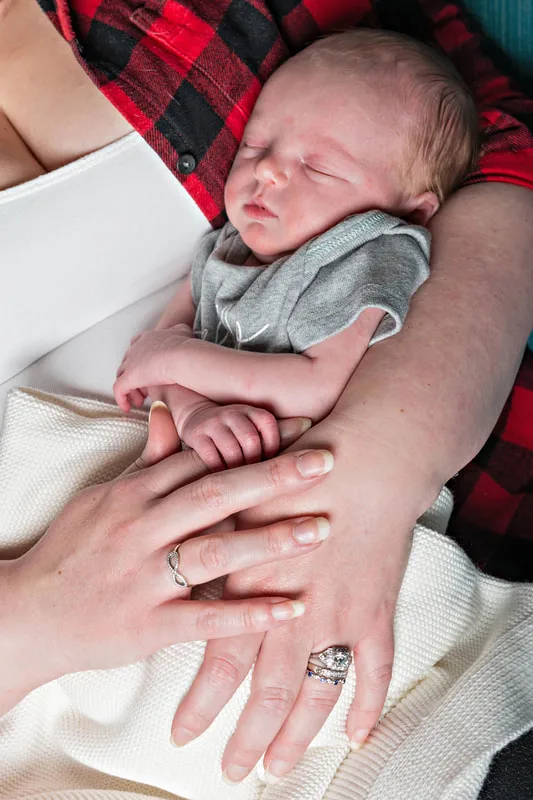 Color close up photo of a newborn baby boy being held in his mother's arms while he holds onto her finger. The photo is by Laura Pea Photography, a Long Island newborn and pet photographer. 