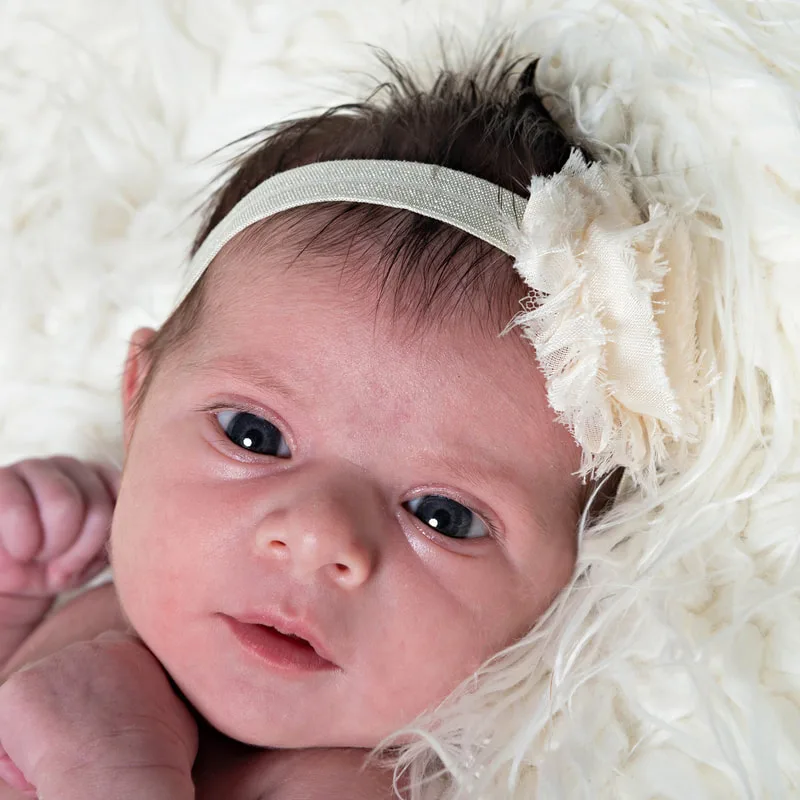 Color closeup photo of a newborn baby girl with a white headband against a white furry rug. The photo was taken by Long Island newborn and pet photographer Laura Pea Photography. 