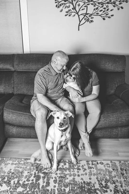 Black and white photo of a newborn baby girl being held by her parents on a couch with their dog on the floor in front of them. The photo was taken by Laura Pea Photography, a Long Island newborn and pet photographer. 