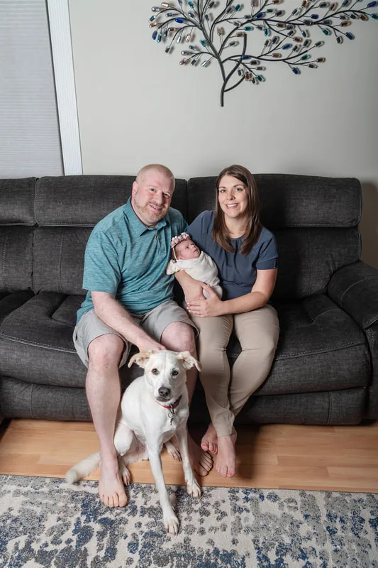 Color photo of a newborn baby girl being held by her parents on a couch with their dog on the floor in front of them. The photo was taken by Laura Pea Photography, a Long Island newborn and pet photographer. 