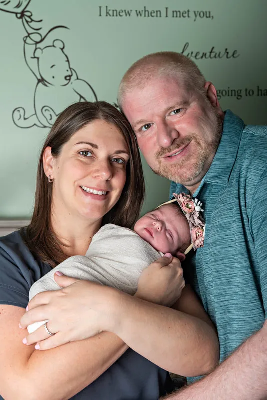 Color photo of a newborn baby girl being held by her parents in her Winnie the Pooh themed nursery. The photo was taken by Laura Pea Photography, a Long Island newborn and pet photographer. 