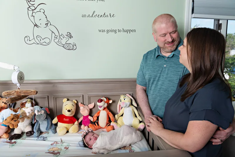 Color photo of a newborn baby girl in her crib with her parents in her Winnie the Pooh themed nursery. The photo was taken by Laura Pea Photography, a Long Island newborn and pet photographer. 