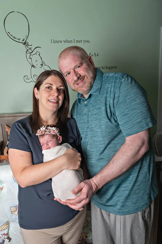 Color photo of a newborn baby girl being held by her parents in her Winnie the Pooh themed nursery. The photo was taken by Laura Pea Photography, a Long Island newborn and pet photographer. 