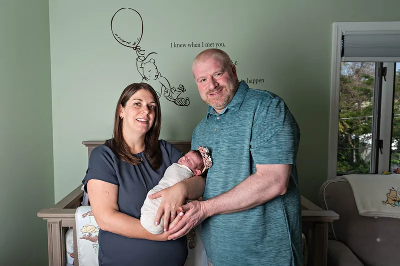 Color photo of a newborn baby girl being held by her parents in her Winnie the Pooh themed nursery. The photo was taken by Laura Pea Photography, a Long Island newborn and pet photographer. 