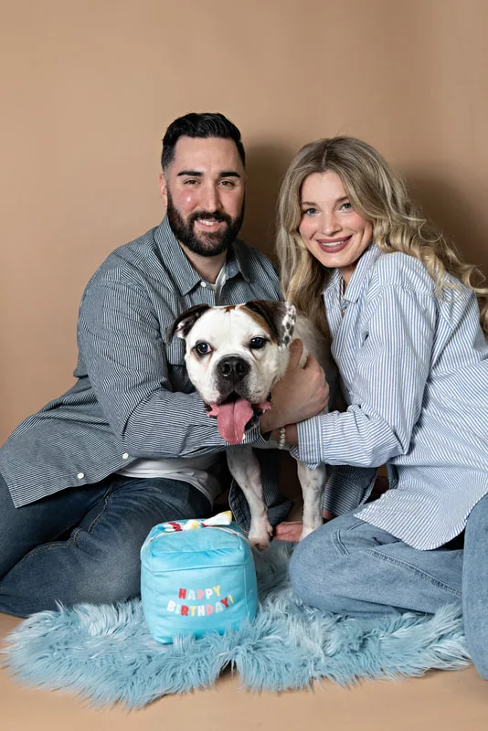 color photo of a bulldog beagle mix dog and his two owners, a white male and female, against a brown backdrop