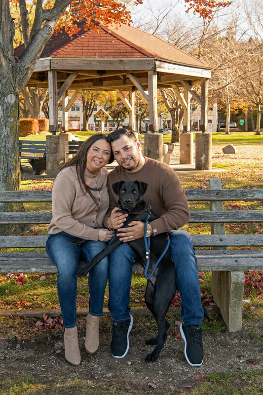 Color photo of a white man and women and their black labrador mix dog. The photo was taken by Laura of Laura Pea Photography, a Long Island based pet photographer. 