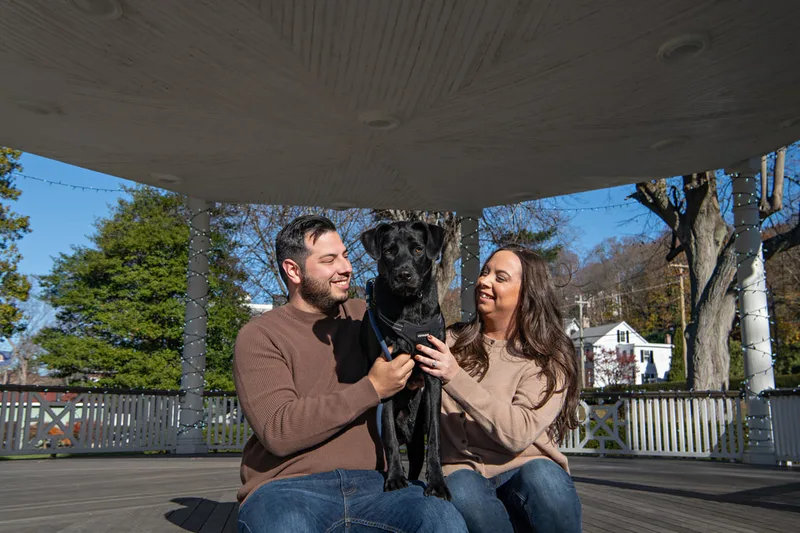 Color photo of a white man and women and their black labrador mix dog. The photo was taken by Laura of Laura Pea Photography, a Long Island based pet photographer. 