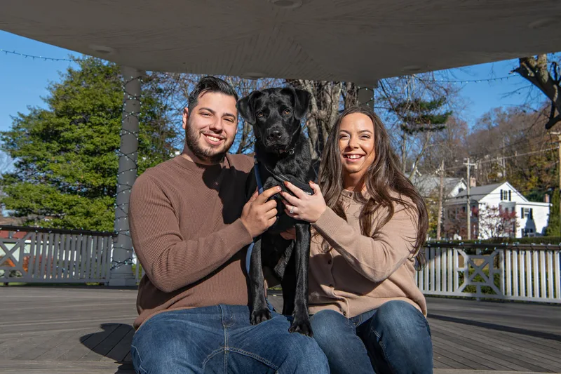Color photo of a white man and women and their black labrador mix dog. The photo was taken by Laura of Laura Pea Photography, a Long Island based pet photographer. 