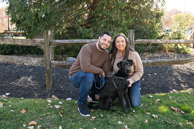 Color photo of a white man and women and their black labrador mix dog. The photo was taken by Laura of Laura Pea Photography, a Long Island based pet photographer. 