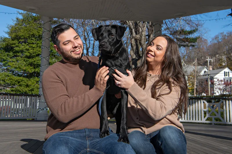 Color photo of a white man and women and their black labrador mix dog. The photo was taken by Laura of Laura Pea Photography, a Long Island based pet photographer. 