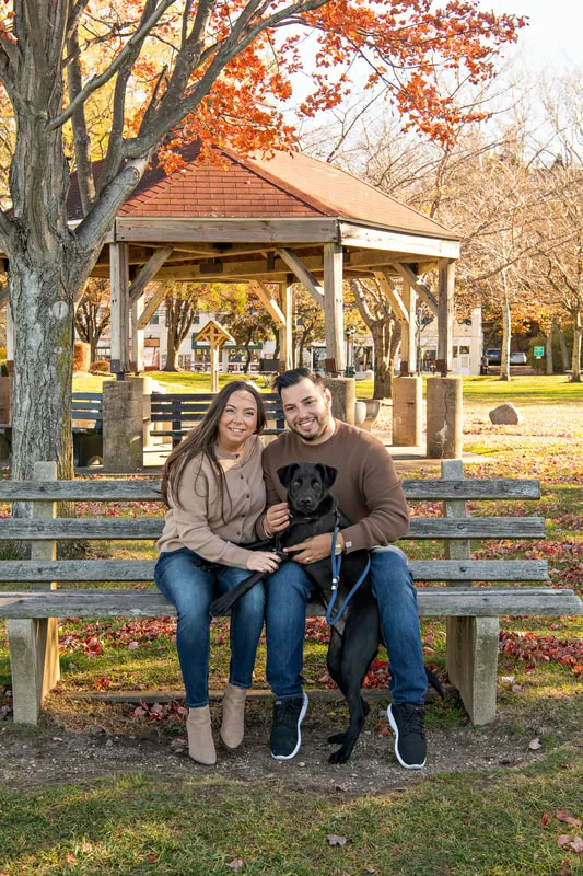 Color photo of a white man and women and their black labrador mix dog. The photo was taken by Laura of Laura Pea Photography, a Long Island based pet photographer. 