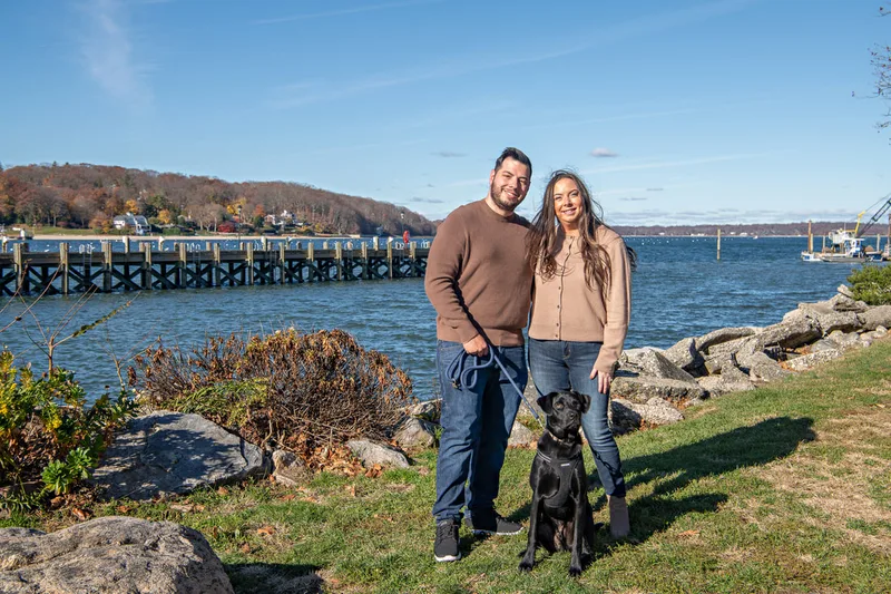 Color photo of a white man and women and their black labrador mix dog. The photo was taken by Laura of Laura Pea Photography, a Long Island based pet photographer. 