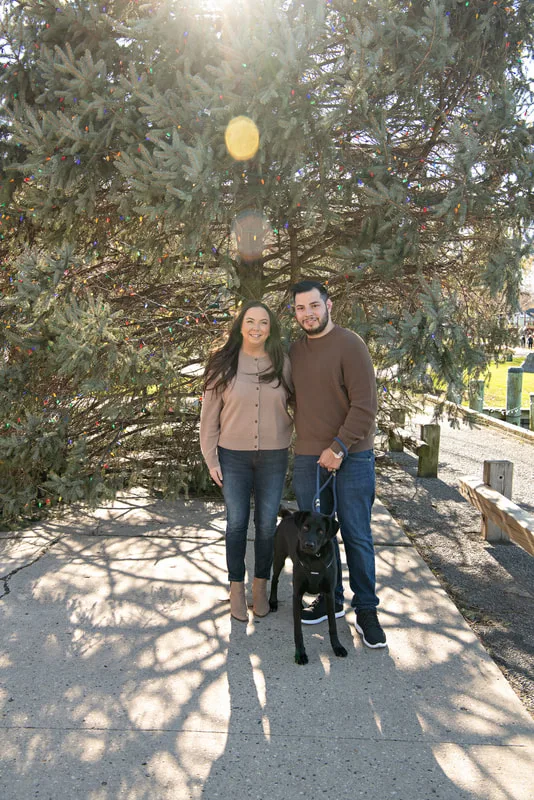 Color photo of a white man and women and their black labrador mix dog. The photo was taken by Laura of Laura Pea Photography, a Long Island based pet photographer. 