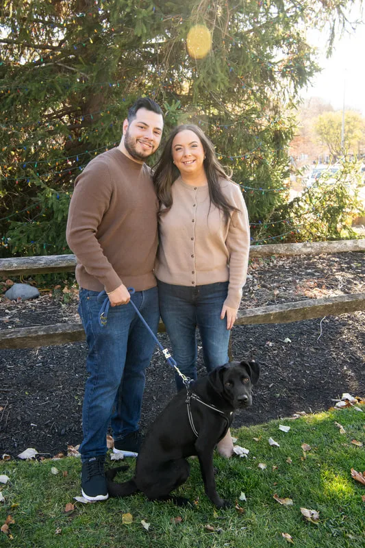 Color photo of a white man and women and their black labrador mix dog. The photo was taken by Laura of Laura Pea Photography, a Long Island based pet photographer. 