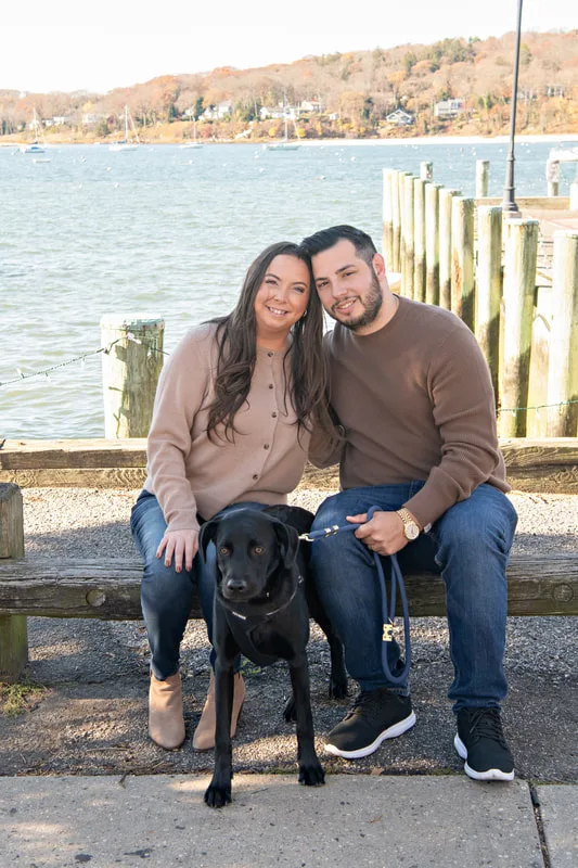 Color photo of a white man and women and their black labrador mix dog. The photo was taken by Laura of Laura Pea Photography, a Long Island based pet photographer. 