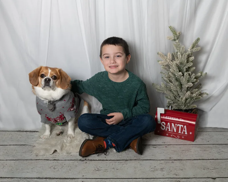 Color photo of a dog and a boy in front of a white winter themed backdrop with pine tree props. The photo was taken at Barkfield Road in East Northport by Laura Paesano of Laura Pea Photography, a Long Island-based pet photographer. 