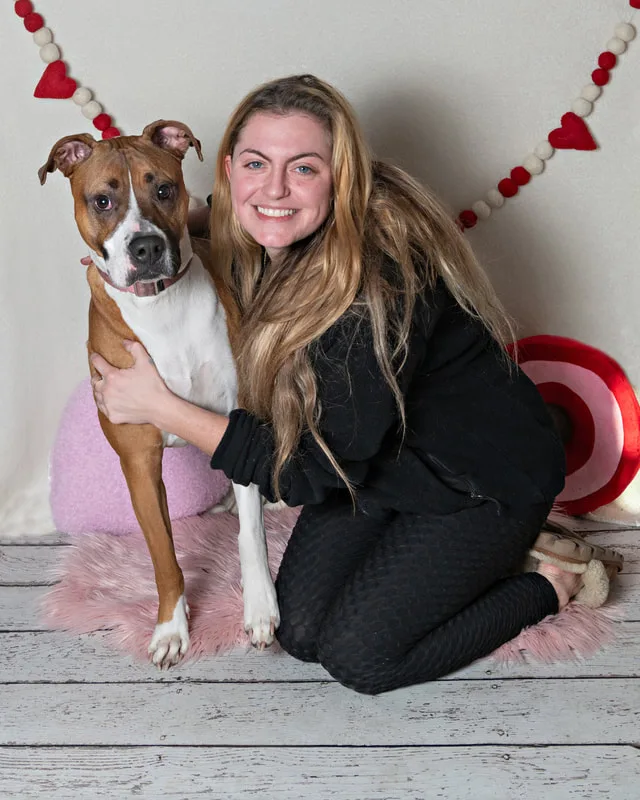 Color photo of a Boxer Pit Bull  mix dog and owner in front of a white backdrop with lots of Valentine's Day props. The photo was taken by Laura Pea Photography, a Long Island pet photographer. 