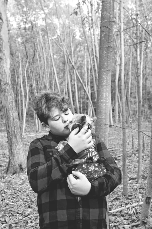 A black and white photo of a Yorkshire "Yorkie" Terrier dog in a red Christmas dress in the woods amongst the fall foliage. She is being held in the arms of her owner, a white teenage boy. The photo is by Laura Pea Photography, a Long Island based pet photographer. 
