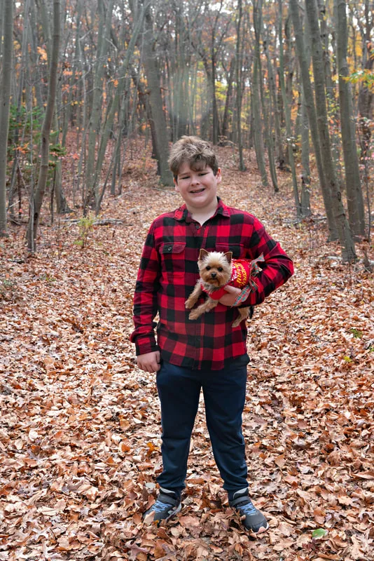 A photo of a Yorkshire "Yorkie" Terrier dog in a red Christmas dress in the woods amongst the fall foliage. She is being held in the arms of her owner, a white teenage boy. The photo is by Laura Pea Photography, a Long Island based pet photographer. 
