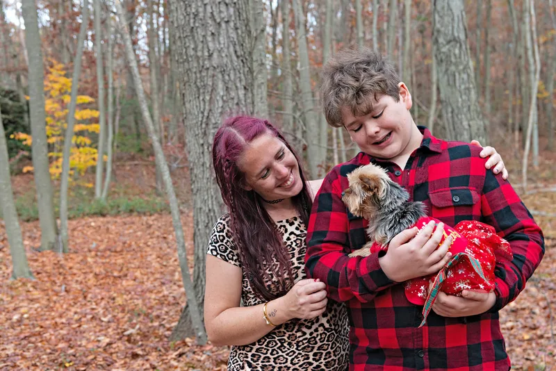 A photo of a Yorkshire "Yorkie" Terrier dog in a red Christmas dress in the woods amongst the fall foliage. She is being held in the arms of her owner, a white teenage boy. They are standing next to the boy's mother, a white woman. Both are looking at the dog. The photo is by Laura Pea Photography, a Long Island based pet photographer. 