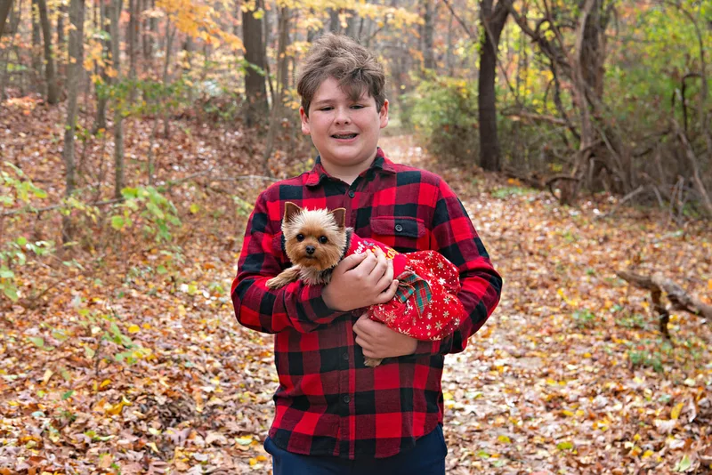 A photo of a Yorkshire "Yorkie" Terrier dog in a red Christmas dress in the woods amongst the fall foliage. She is being held in the arms of her owner, a white teenage boy. The photo is by Laura Pea Photography, a Long Island based pet photographer. 