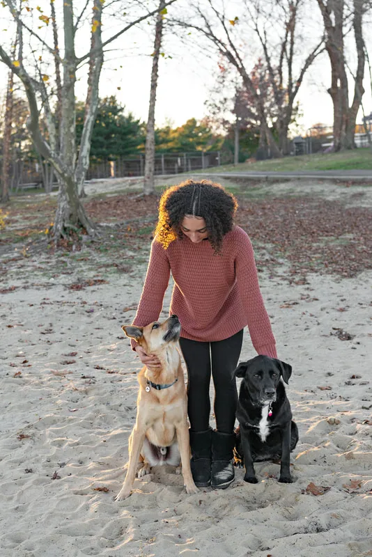 color photo of a teenage girl with a light brown shepherd mix dog and a black and white labrador mix dog sitting on the sandy beach of Lake Ronkonkoma. The girl looks down at her dogs and one of her dogs looks back up at her. Portrait taken by Long Island pet photographer Laura Pea Photography