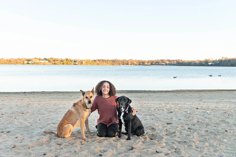 color photo of a teenage girl with a light brown shepherd mix dog and a black and white labrador mix dog sitting on the sandy beach of Lake Ronkonkoma, looking at the camera. Portrait taken by Long Island pet photographer Laura Pea Photography
