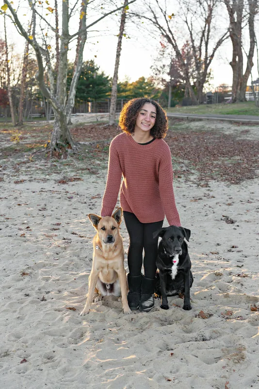 color photo of a teenage girl with a light brown shepherd mix dog and a black and white labrador mix dog standing on the sandy beach of Lake Ronkonkoma, looking at the camera. Portrait taken by Long Island pet photographer Laura Pea Photography