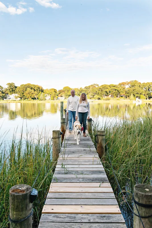 A photo of a brown and white hound mix dog and her two owners, a white man and woman. They are standing on a dock on a creek. The photo is by Laura Pea Photography, a Long Island based pet photographer. 