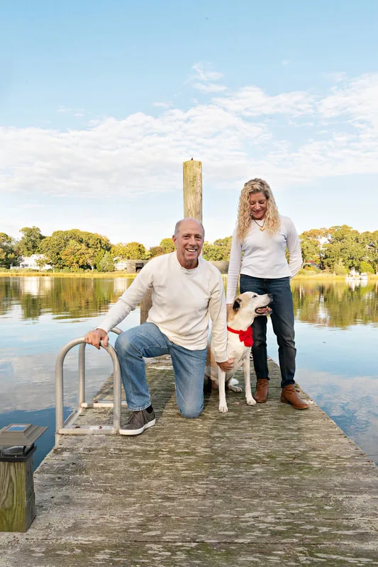 A photo of a brown and white hound mix dog and her two owners, a white man and woman. They are standing on a dock on a creek. The photo is by Laura Pea Photography, a Long Island based pet photographer. 