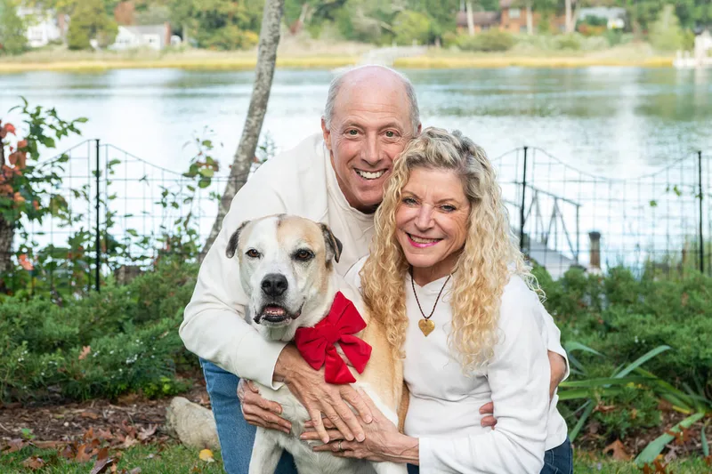 A close up photo of a brown and white hound mix dog and her two owners, a white man and woman.  They are outside in front of a creek. The photo is by Laura Pea Photography, a Long Island based pet photographer. 