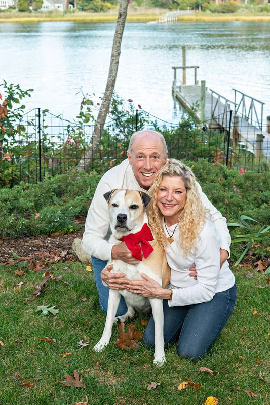 A photo of a brown and white hound mix dog and her two owners, a white man and woman. They are kneeling on the ground outside in front of a creek. The photo is by Laura Pea Photography, a Long Island based pet photographer. 