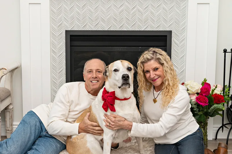 A photo of a brown and white hound mix dog and her two owners, a white man and woman. They are sitting on the floor in front of a fireplace. The photo is by Laura Pea Photography, a Long Island based pet photographer. 