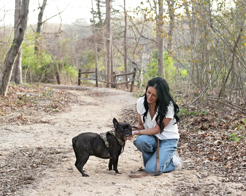 Color photo of a brindle French bulldog dog and his owner, a white woman with black hair wearing a white t-shirt and blue jeans. They pose together in the woods at West Hills Park in Suffolk County. The photo was taken by Laura Pea Photography, a Long Island based pet photographer. 