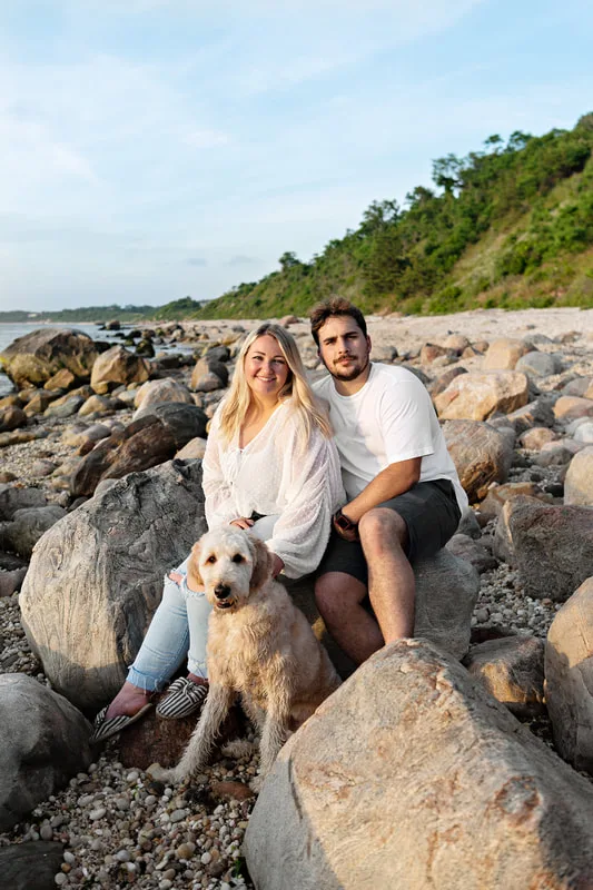 color photo of a golden doodle dog at a beach on Long Island. He's sitting with his owners on a boulder