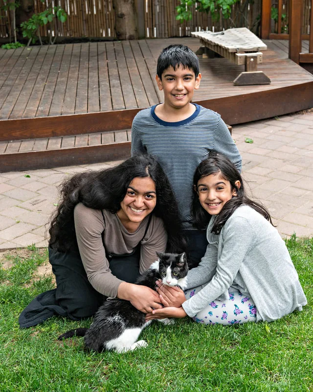 A color photo of a cat with a boy and two girls. The photo was taken by pet photographer Laura Paesano of Laura Pea Photography for a pet portrait program held by the Middle Country Public Library in their outdoor Nature Explorium. 