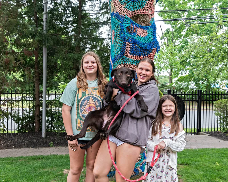 A color photo of a chocolate Labrador dog with a disability being held by three girls. The photo was taken by pet photographer Laura Paesano of Laura Pea Photography for a pet portrait program held by the Middle Country Public Library in their outdoor Nature Explorium. 