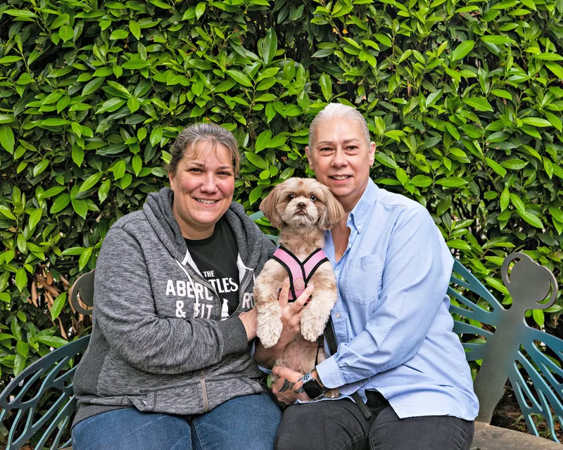 A color photo of a dog with two women sitting on a bench. The photo was taken by pet photographer Laura Paesano of Laura Pea Photography for a pet portrait program held by the Middle Country Public Library in their outdoor Nature Explorium. 