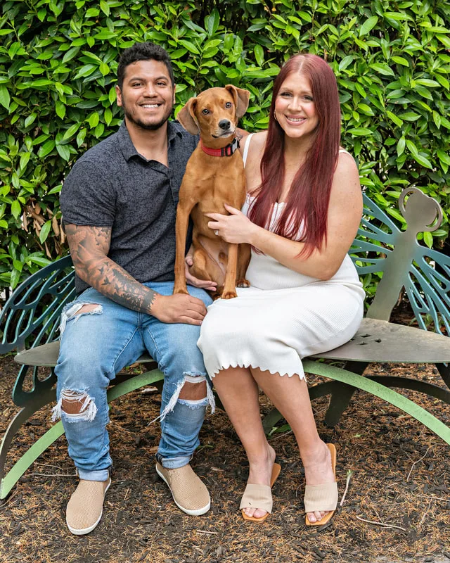 A color photo of a man, woman and dog sitting on a bench. The photo was taken by pet photographer Laura Paesano of Laura Pea Photography for a pet portrait program held by the Middle Country Public Library in their outdoor Nature Explorium. 