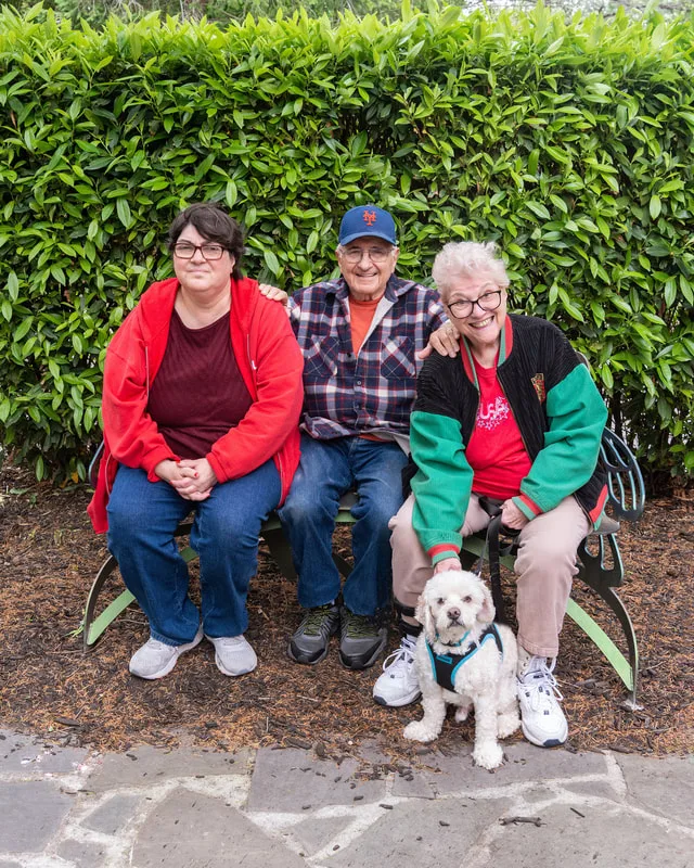 A color photo of a mother, father and daughter with their dog sitting on a bench. The photo was taken by pet photographer Laura Paesano of Laura Pea Photography for a pet portrait program held by the Middle Country Public Library in their outdoor Nature Explorium. 