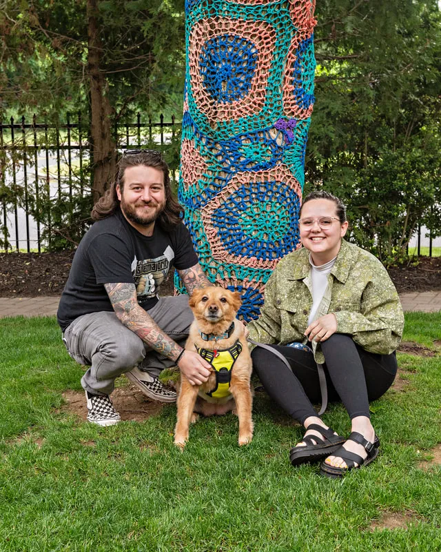 A color photo of a man, woman and their dog kneeling on the ground. The photo was taken by pet photographer Laura Paesano of Laura Pea Photography for a pet portrait program held by the Middle Country Public Library in their outdoor Nature Explorium. 
